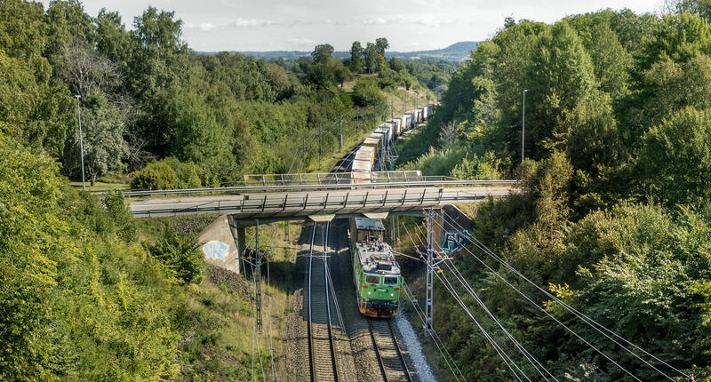 Godståg åker genom ett grönt och mulligt landskap. När bilden tas passerar godsloket under en bro. Solig höstdag ger ett mysigt intryck av bilden. 
