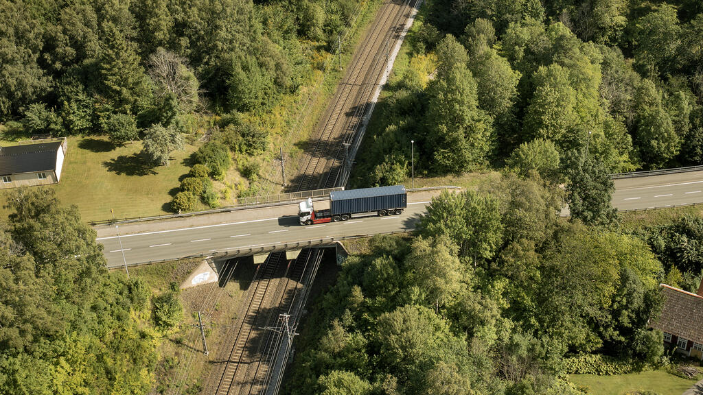 En lastbil med containersläp korsar järnväg på en bro i ett skogsnära område.